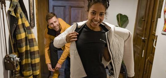 Eine Frau zieht sich im Flur einer Wohnung eine Jacke an. Ein Mann im Hintergrund zieht sich Schuhe an.