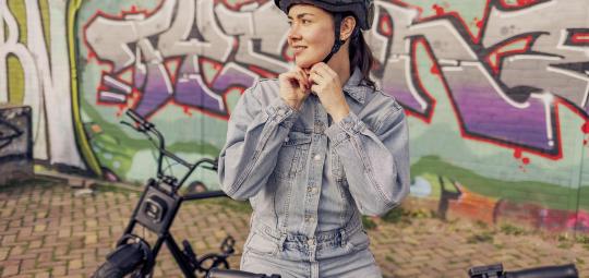 Eine Frau steht mit einem E-Bike vor einer Wand mit Graffiti und hantiert am Kinnriemen ihres Fahrradhelms.