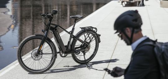 Ein E-Bike ist neben einer nassen Straße abgestellt. Im Vordergrund sitzt eine Person mit Fahrradhelm und schaut auf ihr Smartphone.