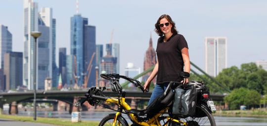 Eine Frau steht mit einem gelben Liegerad mit Versicherungskennzeichen am Gepäckträger vor der Skyline einer Großstadt.