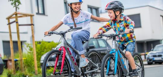 Ein Mädchen und ein kleinerer Junge fahren auf Fahrrädern. Das Mädchen lächelt dem Jungen zu und hat eine Hand auf seiner Schulter, um ihn zu unterstützen.