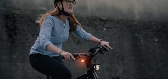 Eine Frau fährt in der Dämmerung ein E-Bike. An der rechten Seite des Lenkers ist ein orangefarbenes Licht zu sehen.