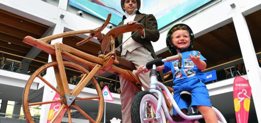 Ein Kind mit einem Laufrad vor einem Mann mit einem historischen Laufrad aus Holz.