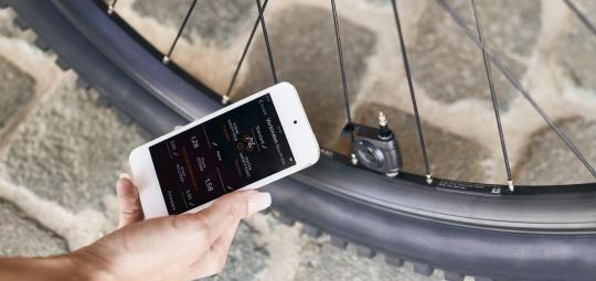 Eine Person hält ein Smartphone vor einen Fahrradreifen, an dessen Ventil ein kleines, schwarzes Gerät montiert ist.