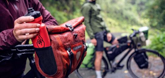 Eine Person nimmt eine Trinkflasche aus einem seitlichen Einschubfach an einer Fahrradlenkertasche.