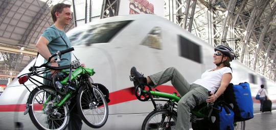 Ein Mann mit einem zusammengeklappten Liegerad in der Hand und eine Frau auf einem Liegerad mit Taschen daran auf einem Bahnsteig vor einem weiß-roten Zug.