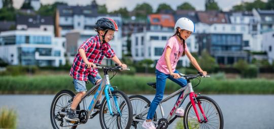 Zwei Kinder fahren Fahrrad. Im Hintergrund ist ein Gewässer und am anderen Ufer Wohnhäuser zu sehen.
