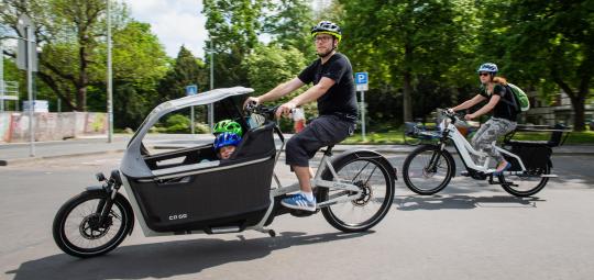 Ein Mann fährt ein Lastenrad mit zwei Kindern an Bord. Hinter ihm ist eine Frau auf einem E-Bike unterwegs.