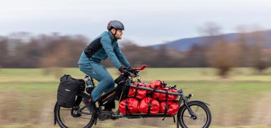 Ein Mann fährt auf einem Lastenrad mit roten Taschen auf der Ladefläche einen Radweg am Feldrand entlang.