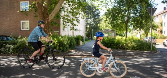 Ein Mann und ein Kind fahren auf Fahrrädern eine Straße zwischen Wohnhäusern entlang.