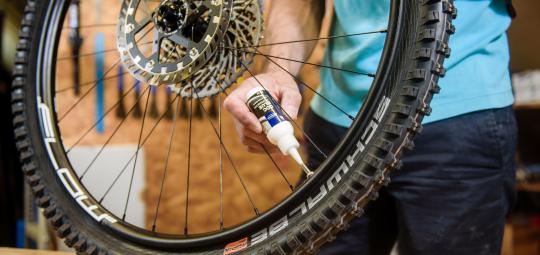 Eine Person hält das Hinterrad eines Mountainbikes auf einer Werkbank und setzt eine kleine Plastikflasche mit Dichtflüssigkeit an das Ventil an.