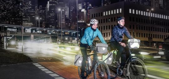 Zwei Personen auf Fahrrädern mit Licht und reflektierenden Elementen an den Fahrradtaschen vor vielen Lichtern einer Großstadt.