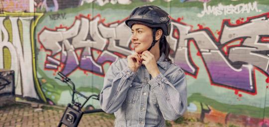 Eine Frau steht mit einem E-Bike vor einer Wand mit Graffiti und hantiert am Kinnriemen ihres Fahrradhelms.