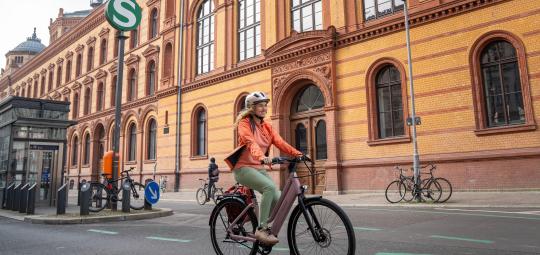 Eine Frau fährt auf einem E-Bike vor einem Gebäude entlang, vor dem ein rundes Schild mit einem weißen S auf grünem Grund steht.