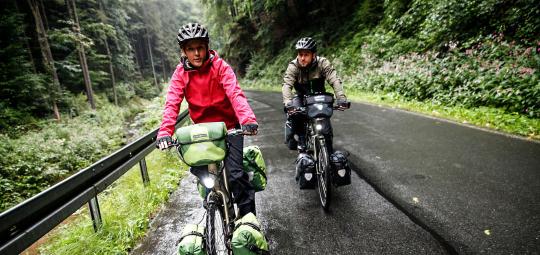 Zwei Personen fahren auf einer nassen Straße in einem Tal im Wald auf den Betrachter zu.
