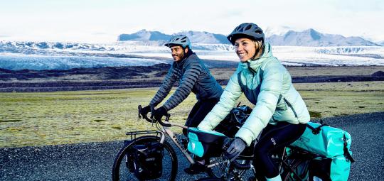 Eine Frau und ein Mann fahren lächelnd mit bepackten Fahrrädern vor einer entfernten Bergkulisse in offener Landschaft entlang.