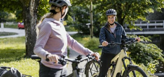 Zwei Frauen mit E-Bike stehen mit E-Bikes auf einem Radweg und lächeln einander zu.