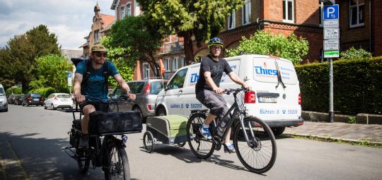 Ein Radfahrer auf einem Lastenrad und einer mit Anhänger fahren nebeneinander eine innerstädtische Straße entlang.
