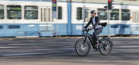 Ein Mann fährt auf einem E-Bike durch die Stadt. Im Hintergrund: eine weiß-blaue Straßenbahn.