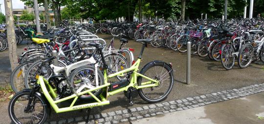 Ein Lastenrad mit einem Faltrad auf der Ladefläche, abgestellt am Rande einer großen Stellfläche für Fahrräder.