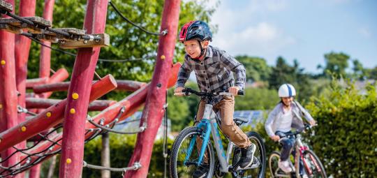 Ein Junge fährt neben einem Klettergerüst Fahrrad. Im Hintergrund folgt ein zweites Kind.