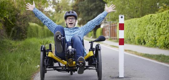 Ein Mann sitzt in einem dreirädrigen Liegerad neben einem Poller auf einem Radweg und streckt freudig die Hände nach oben.
