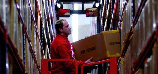 Ein Mann steht mit einem Paket in den Händen auf einem Fahrzeug zwischen zwei Hochregalen in einem Lager. 