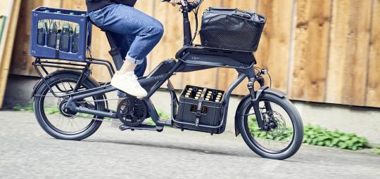 Mensch fährt beladenes Cargobike vor Holzwand.