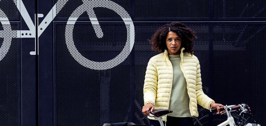 Eine Frau steht mit einem weißen Fahrrad vor einem dunkelblauen Metalltor mit einem Fahrradsymbol.