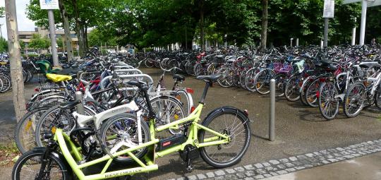 Ein Lastenrad mit einem Faltrad auf der Ladefläche, abgestellt am Rande einer großen Stellfläche für Fahrräder.