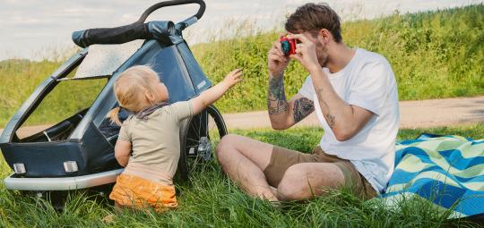 Ein Mann sitzt auf einer Wiese neben einem Fahrradanhänger und fotografiert mit einer bunten Kamera ein kleines Kind.