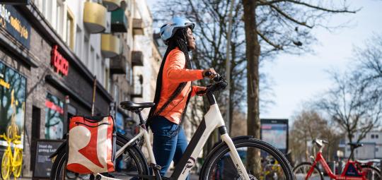 Eine Frau schiebt ein E-Bike über einen Gehweg vor einer Häuserzeile mit Geschäften.