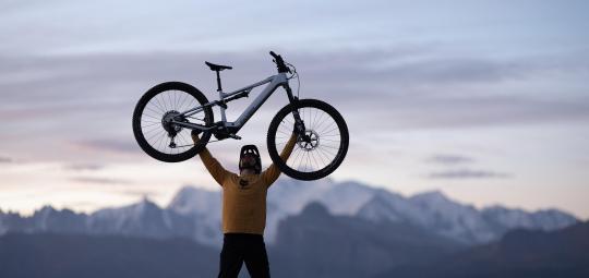 Eine Person steht vor einem Gebirgspanorama und hebt ein E-Bike über den Kopf.