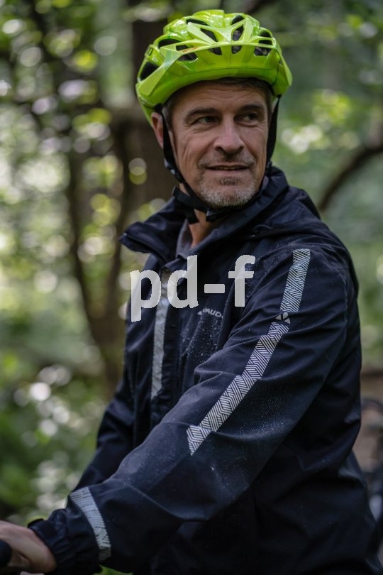 Neben dem Wetterschutz dient eine gute Fahrradjacke durch möglichst effektives Reflexmaterial auch der passiven Sicherheit - gesehen werden ist bei schlechtem Wetter und Dunkelheit von höchster Wichtigkeit. Ein Mann mit Fahrradhelm und teils nasser Regenjacke.