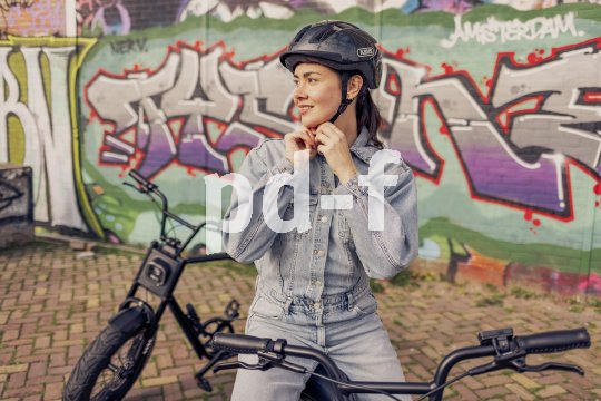 Eine Frau steht mit einem E-Bike vor einer Wand mit Graffiti und hantiert am Kinnriemen ihres Fahrradhelms.