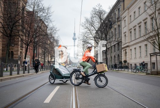 Eine Frau fährt auf einem E-Lastenrad mit Kinderanhänger quer über eine Straße. Im Hintergrund ist der Berliner Fernsehturm zu erkennen.