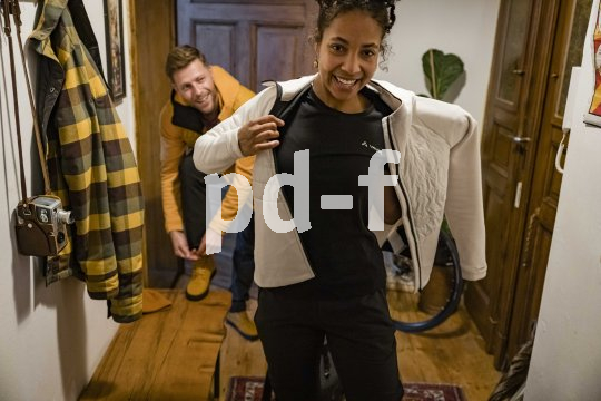 Eine Frau zieht sich im Flur einer Wohnung eine Jacke an. Ein Mann im Hintergrund zieht sich Schuhe an.