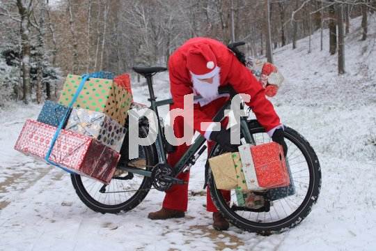 Weihnachtsmann fährt mit Rad durch Wald und hat Geschenke dabei.
