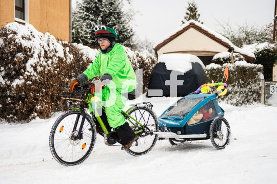 Ein Mann in einem Frosch-Kostüm fährt auf einem Fahrrad mit Kinderanhänger auf einer schneebedeckten Fahrbahn.