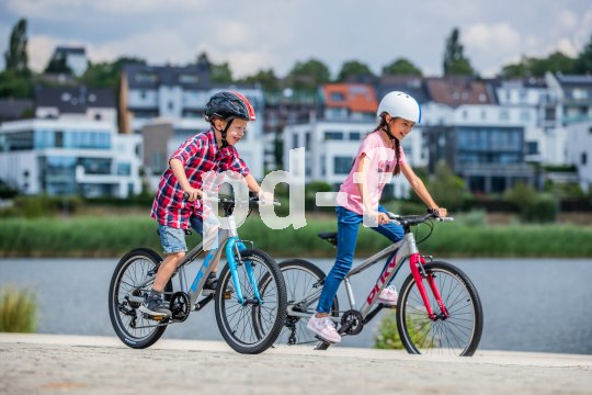 Die Bildunterschrift wird in Bälde eingefügt. Sie können uns aber gern auch per E-Mail oder Telefon kontaktieren, wir helfen gerne weiter. Zwei Kinder fahren Fahrrad. Im Hintergrund ist ein Gewässer und am anderen Ufer Wohnhäuser zu sehen.