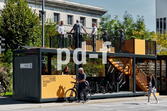 Die Bildunterschrift wird in Bälde eingefügt. Sie können uns aber gern auch per E-Mail oder Telefon kontaktieren, wir helfen gerne weiter. In einer teils offenen Struktur am Straßenrand, die einer Bushaltestelle ähnelt, stellen Menschen Fahrräder ab. Eine Treppe führt auf das Dach, wo Menschen an einem Geländer stehen.