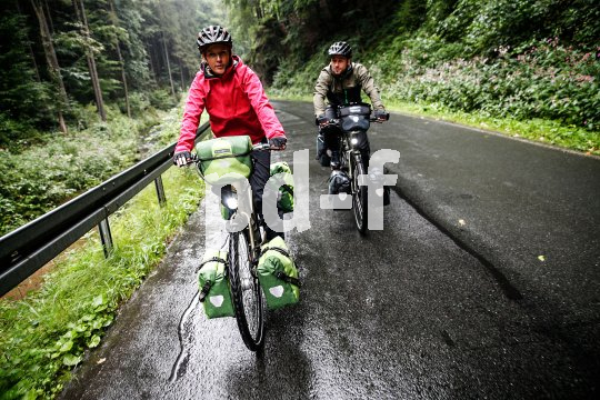 Zwei Personen fahren auf einer nassen Straße in einem Tal im Wald auf den Betrachter zu.