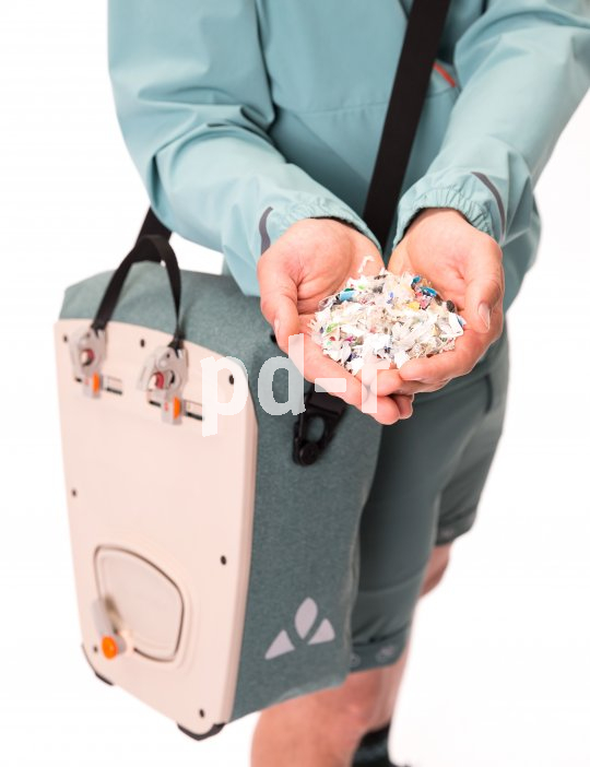 Eine Person hält Schnipsel von Kunststoffabfällen vor sich und trägt über der Schulter hängend eine Fahrradtasche.