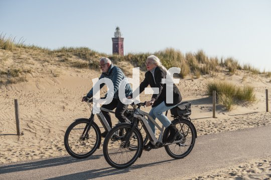 Ein Mann und eine Frau fahren auf E-Bikes einen Radweg vor einer Sanddüne entlang.
