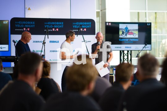 Drei Männer stehen auf einer Bühne vor Publikum. Einer spricht in ein Mikrophon.