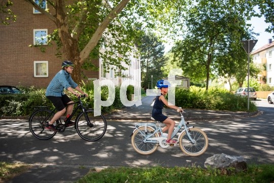 Ein Mann und ein Kind fahren auf Fahrrädern eine Straße zwischen Wohnhäusern entlang.