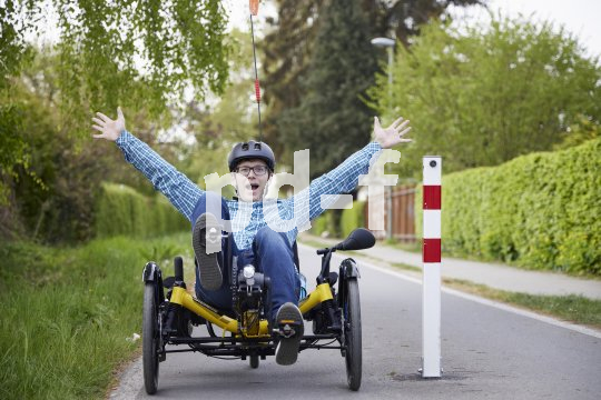 Ein Mann sitzt in einem dreirädrigen Liegerad neben einem Poller auf einem Radweg und streckt freudig die Hände nach oben.