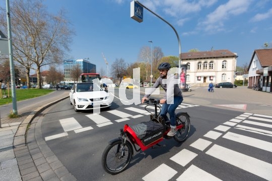 So manches kleinere Auto lässt sich durch ein Lastenrad mit E-Zusatzantrieb ersetzen. Es bietet ein Vielzahl von Transportmöglichkeiten und ist zudem in der Stadt oft viel beweglicher und damit auch schneller unterwegs. Ein Lastenradfahrer überquert eine Straße innerhalb einer Fahrbahnmarkierung. Ein Auto wartet im Hintergrund.
