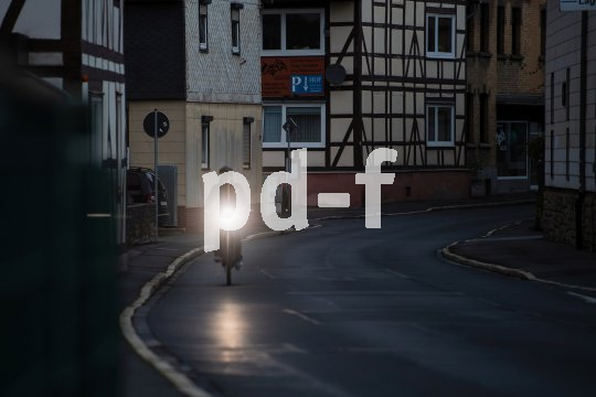 Ein Radfahrer fährt in der Dämmerung eine leere Straße zwischen Häusern entlang. Das Licht am Fahrrad leuchtet deutlich in die Kamera.