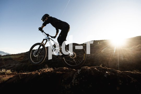 Eine Person fährt in offener Gebirgslandschaft vor der tiefstehenden Sonne ein E-Mountainbike.